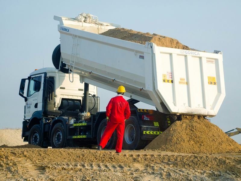 A tipper truck unloading sand while a worker in a red overall supervises