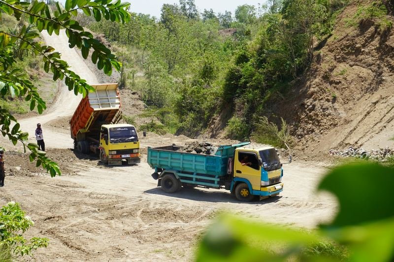 Two tipper trucks on a dirt road cut through green hills